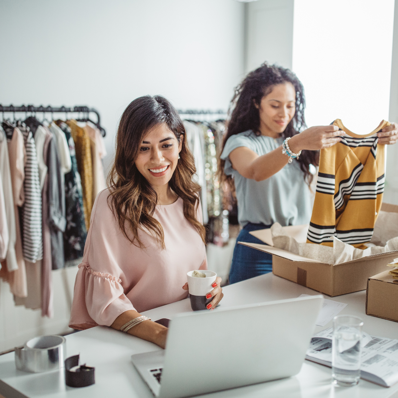 A clothing store owner is checking online orders on a laptop, while a staff member nearby is organizing clothes in the store.