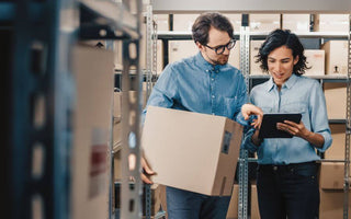 Two staffs reviewing inventory in a stockroom with a tablet and a box of goods.