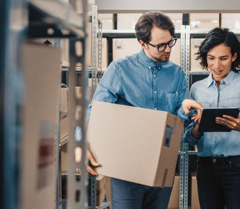 Two staffs reviewing inventory in a stockroom with a tablet and a box of goods.