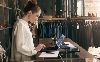 Clothing store owner reviewing online orders on a laptop at the checkout counter.