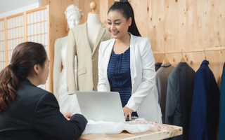 Fashion retailer checking online sales data on a laptop while interacting with a customer.