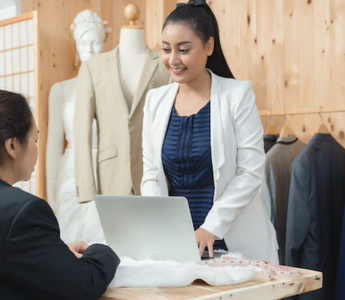 Fashion retailer checking online sales data on a laptop while interacting with a customer.